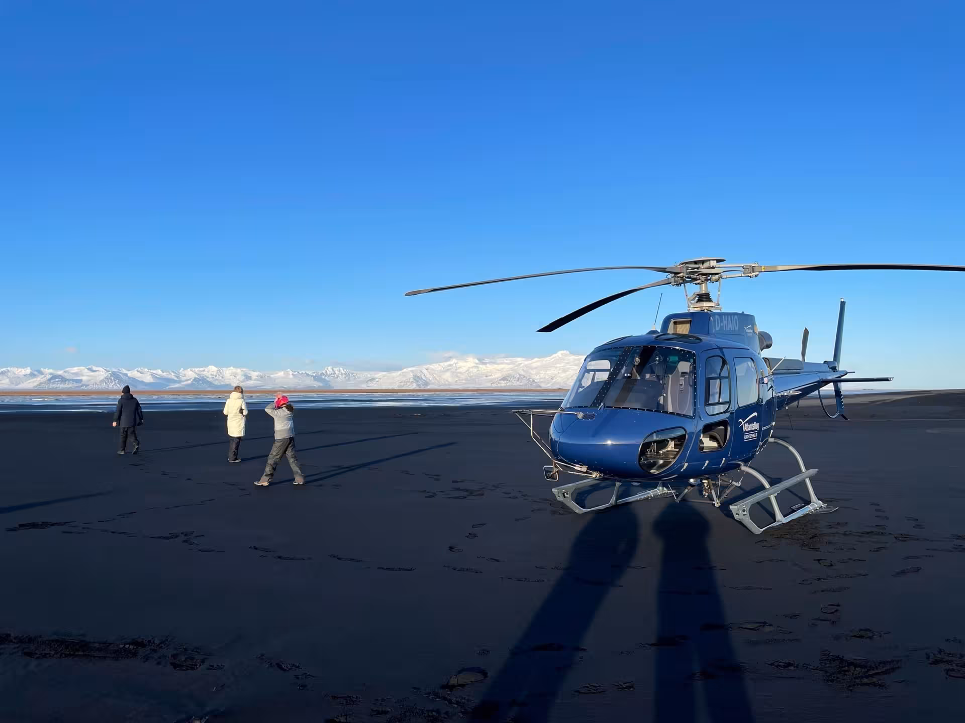 Blue helicopter landed on black sand plains with three tourists and snow-capped mountains in background, Iceland
