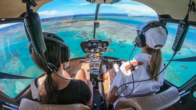 Aerial helicopter cockpit view over turquoise coral reef waters with two passengers wearing headsets