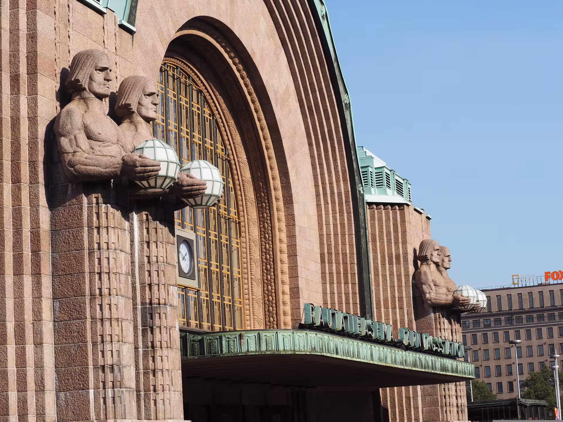 Iconic stone figures and grand entrance of historic Helsinki Central Station under clear blue skies.