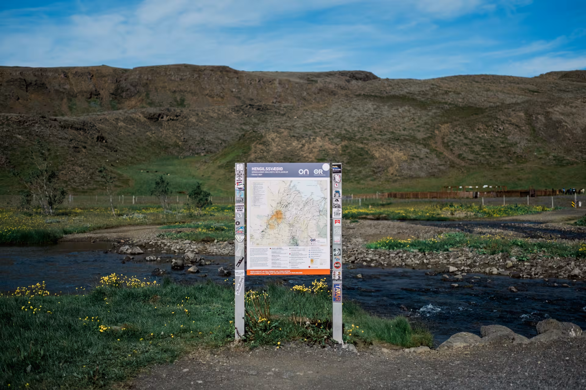 Trail map sign for Hengilssvæðið geothermal area beside a rocky river with green hills in Iceland