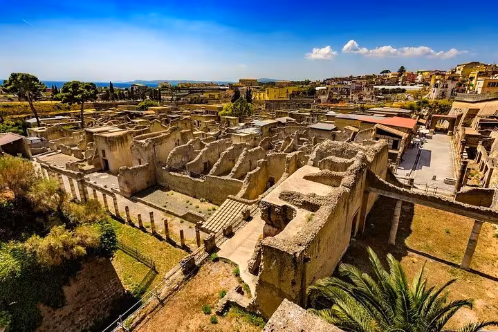 Discover the panoramic view of Hercolaneum's intricate ruins, surrounded by lush greenery and stunning coastal backdrop.