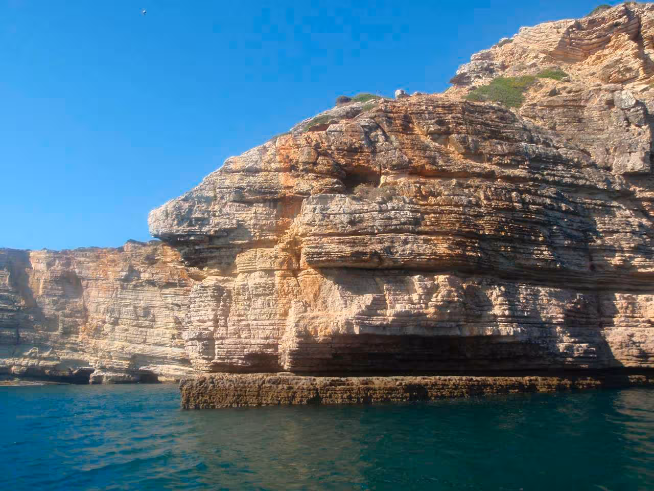 Majestic layered rock formations of a hidden coastal cave under a bright blue sky.