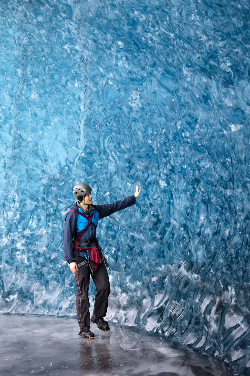 A hiker in climbing gear touches the stunning blue ice wall inside an Iceland glacier cave tour.