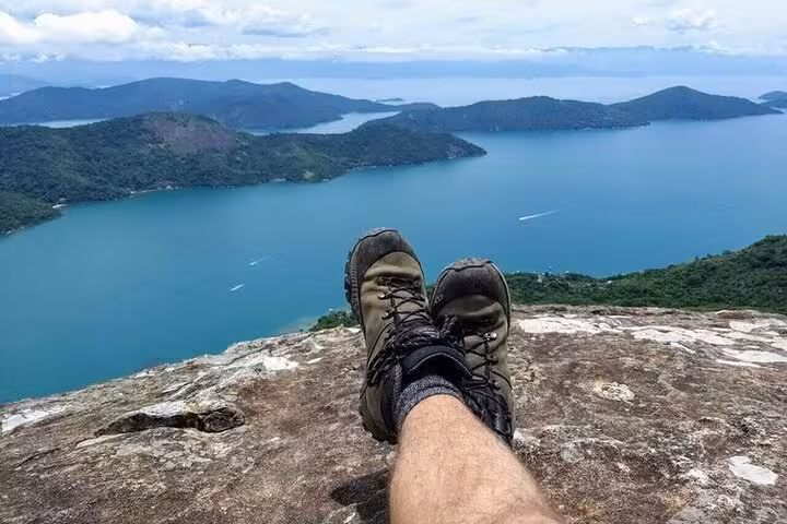 Hiker's boots rest on rocky cliff at Peak Sugar Bread, offering panoramic views of vibrant blue bay and green isles.