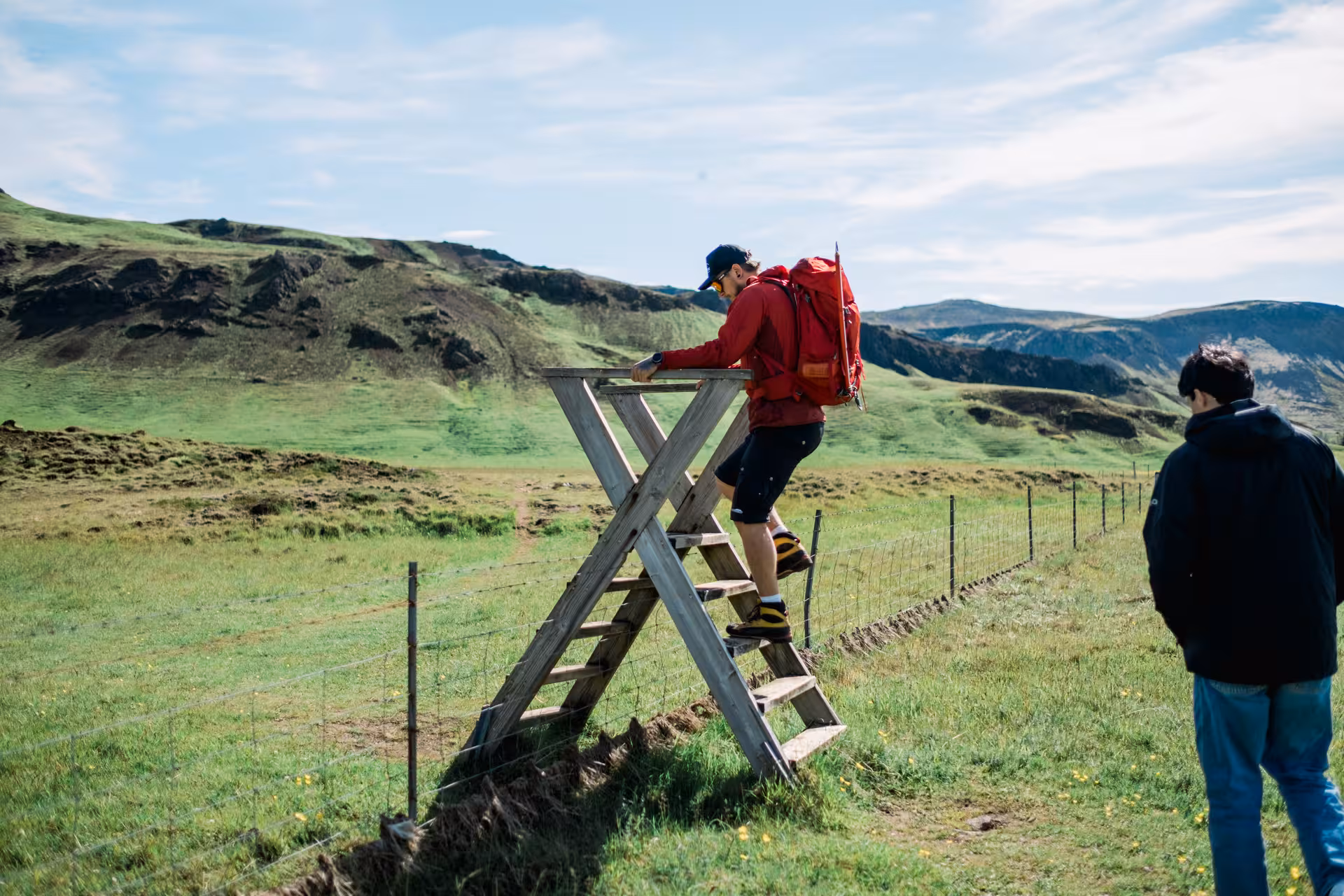 Hiker with red backpack climbing wooden stile over fence in Iceland's green volcanic highlands