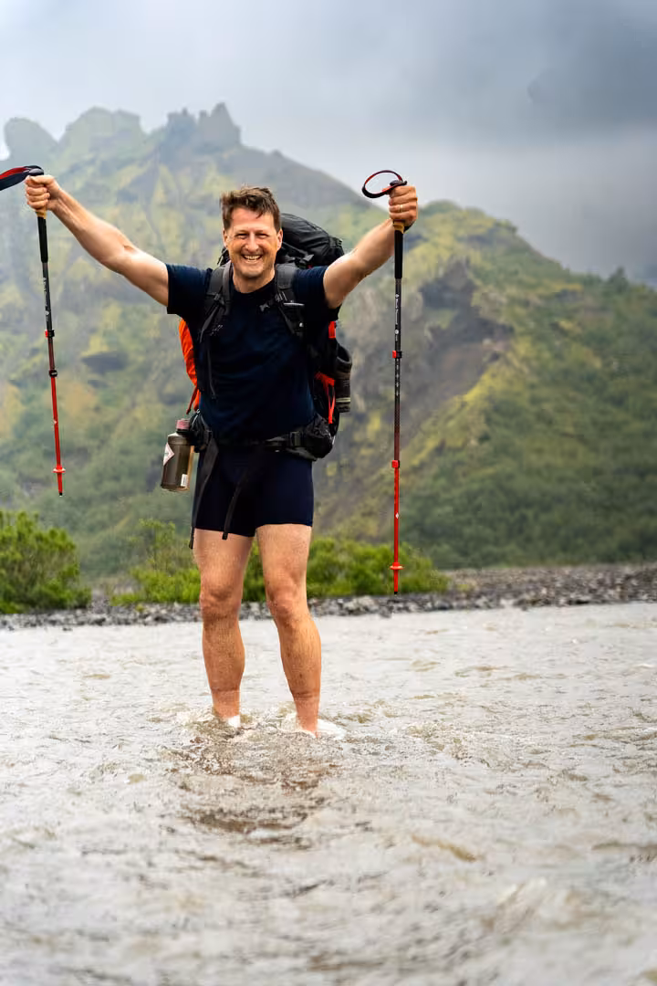 Smiling hiker crossing glacial river with trekking poles raised, green Icelandic mountains in background