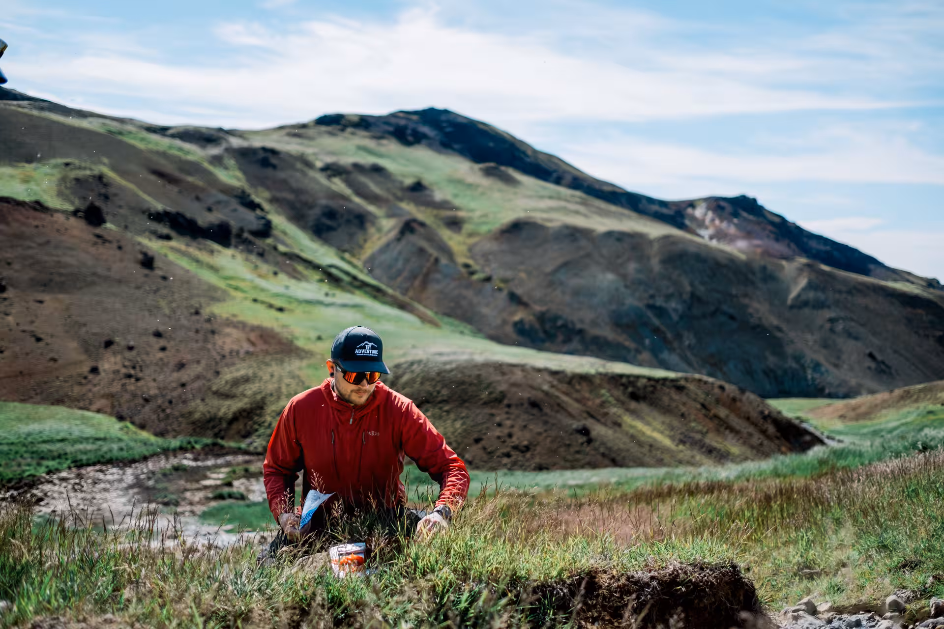 Hiker in red jacket crouching in geothermal valley near Hengill Volcano, Iceland, with green volcanic hills behind