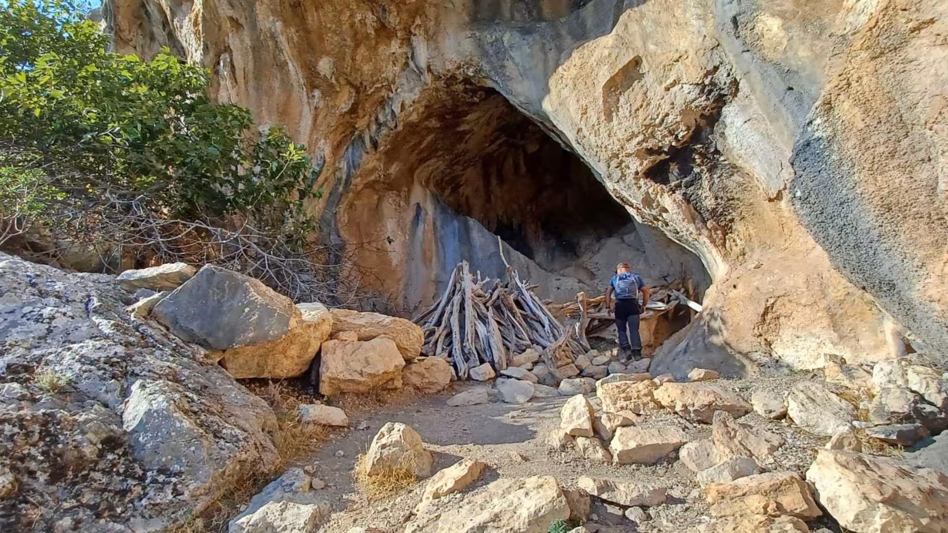 Hiker exploring rugged Gorropu Canyon cave entrance amidst rocky terrain and lush greenery.