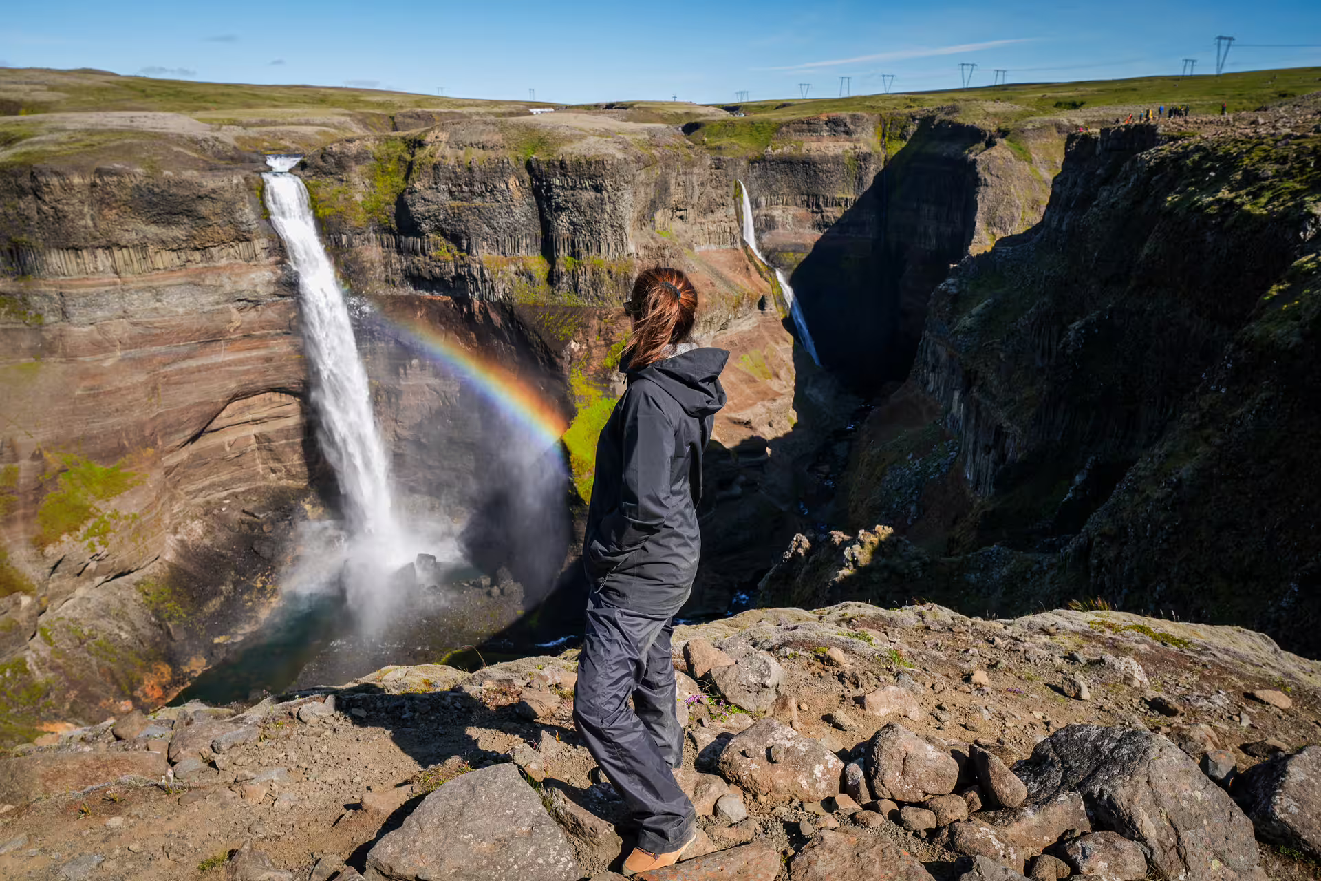 A hiker stands on rocky cliffs overlooking Haifoss waterfall with a vivid rainbow in Iceland's Hidden Highlands tour