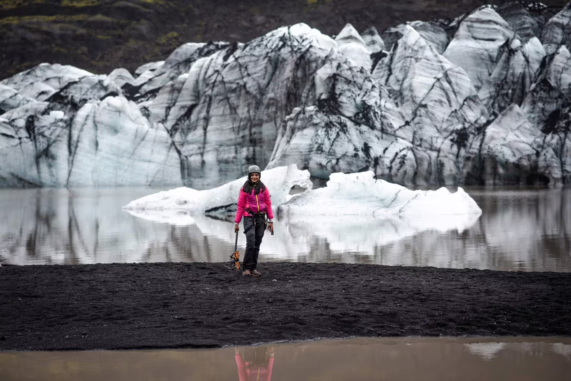 A smiling hiker in a pink jacket holds an ice axe on Iceland's black sand beach with dramatic glacier formations and icebergs on the South Coast private tour.