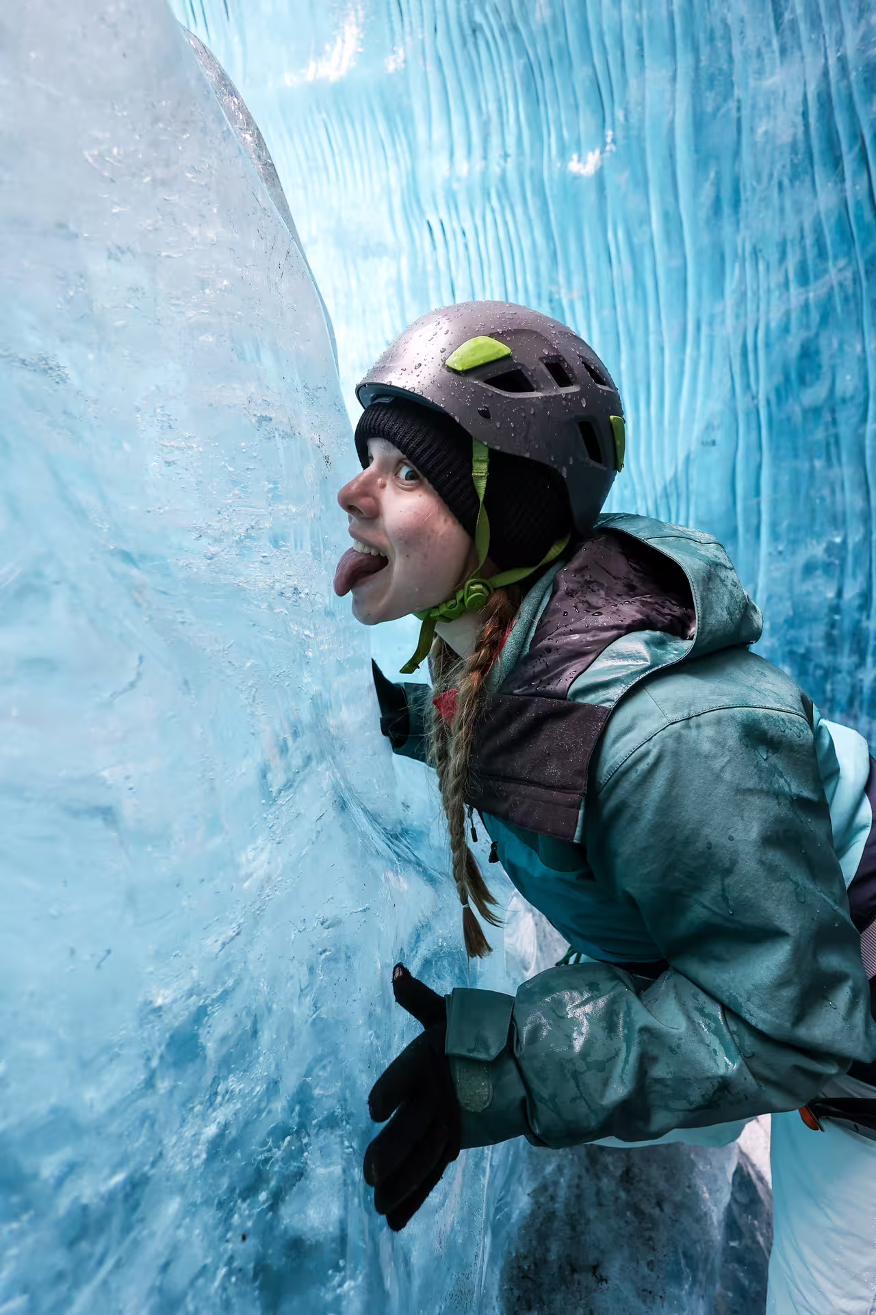 A playful hiker in a helmet and teal jacket licks glacier ice wall during a private glacier hike experience