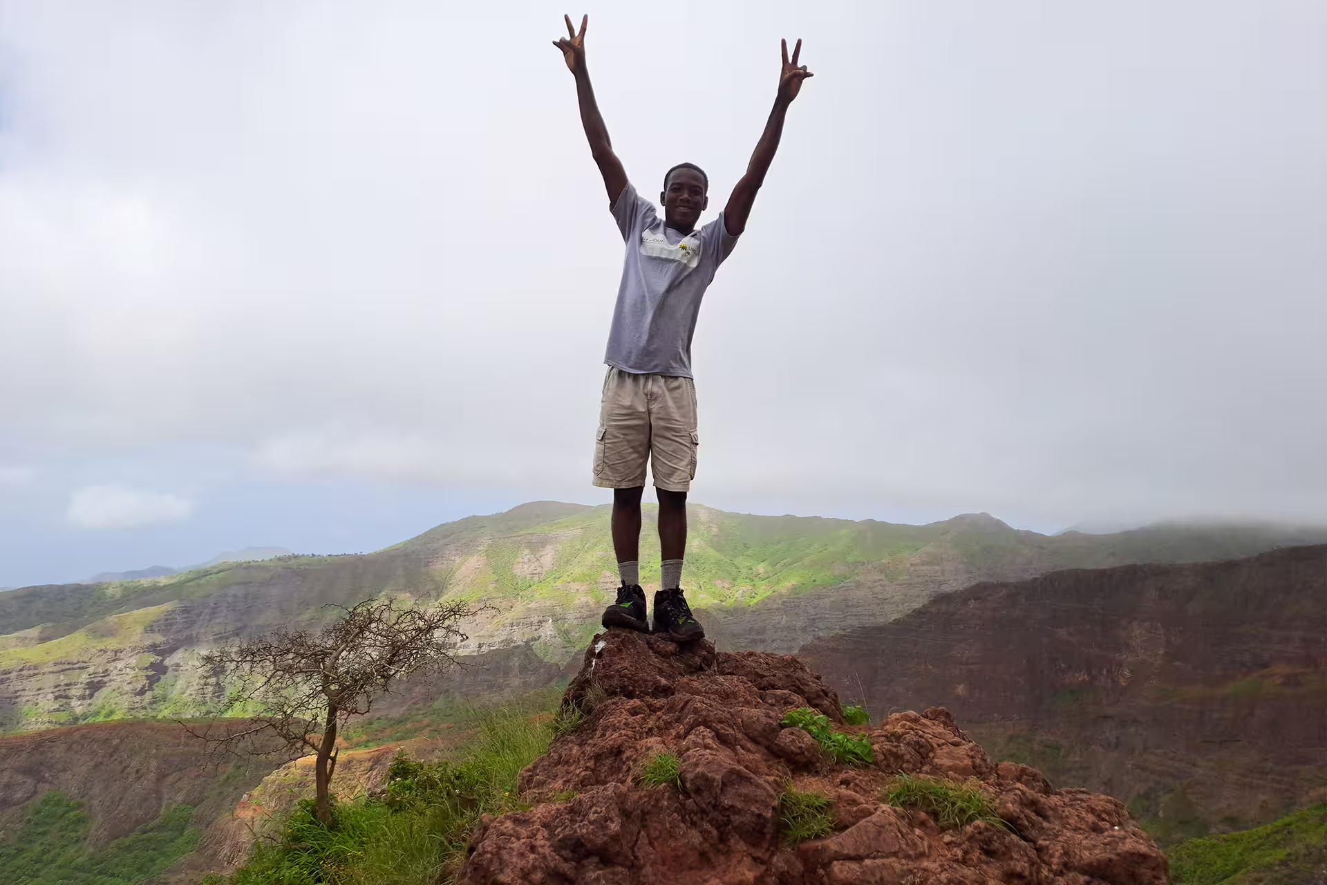 Hiker triumphantly atop rocky summit in Monte Tchota Natural Park, with lush green valleys and misty peaks in view.