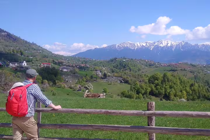 Hiker with red backpack overlooking green valley and snow-capped Piatra Craiului mountains, Romania