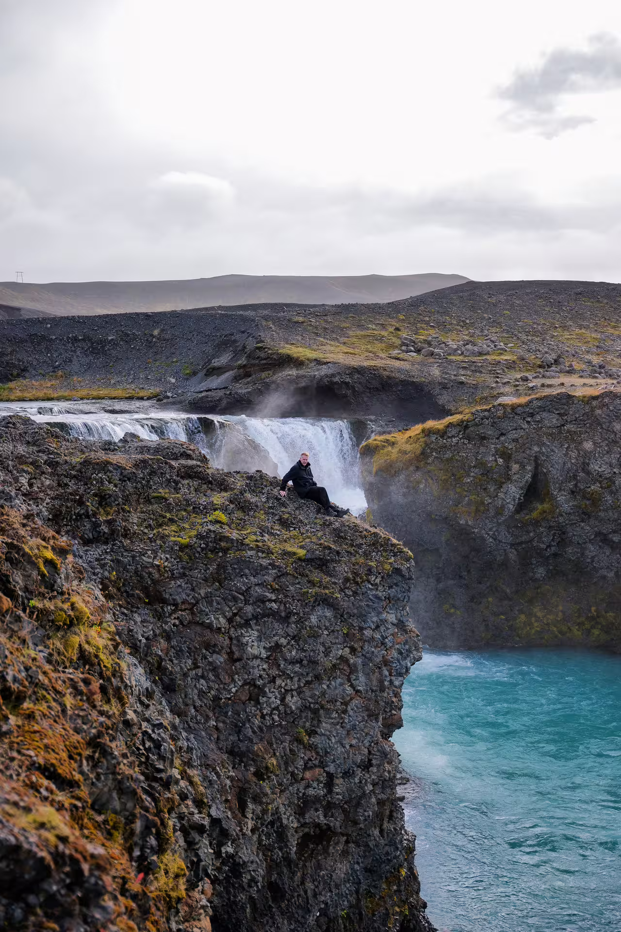 A hiker sits on a mossy volcanic cliff edge beside a roaring Iceland highland waterfall with turquoise glacial river below.