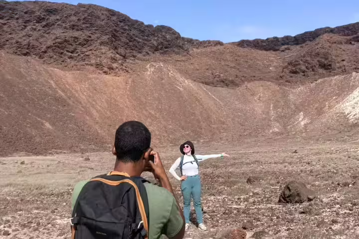 A hiker captures a photo of a friend posing against the rugged volcanic landscape of Volcano Viana, São Vicente.