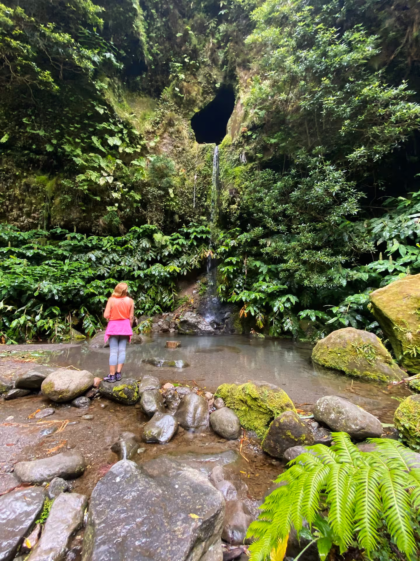 Hiker at Window to Hell waterfall and thermal pool on São Miguel, Azores, surrounded by mossy rocks and vibrant jungle plants