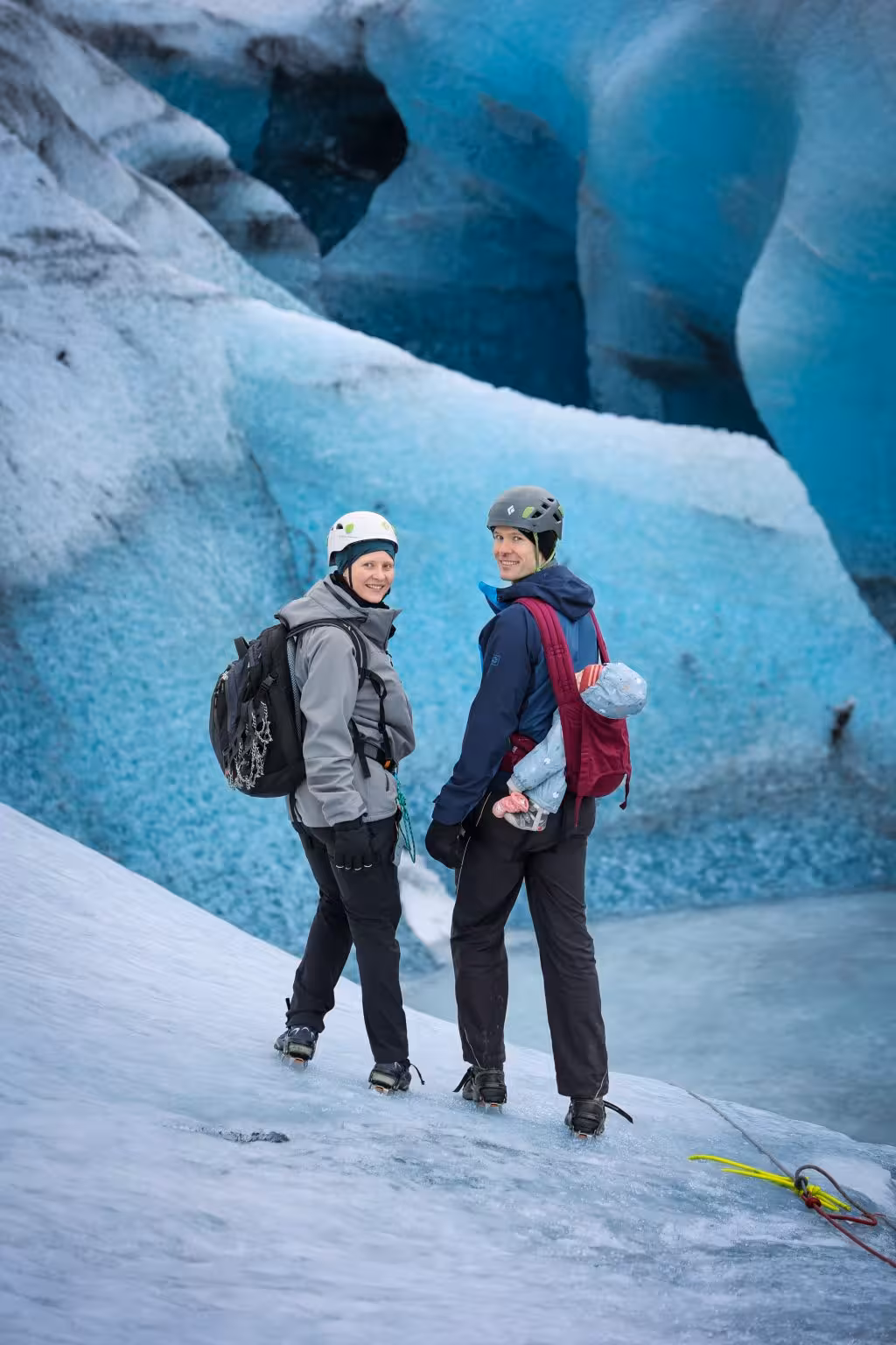 Two hikers with helmets and backpacks standing on an Iceland glacier surrounded by dramatic blue ice walls during a full-day tour.