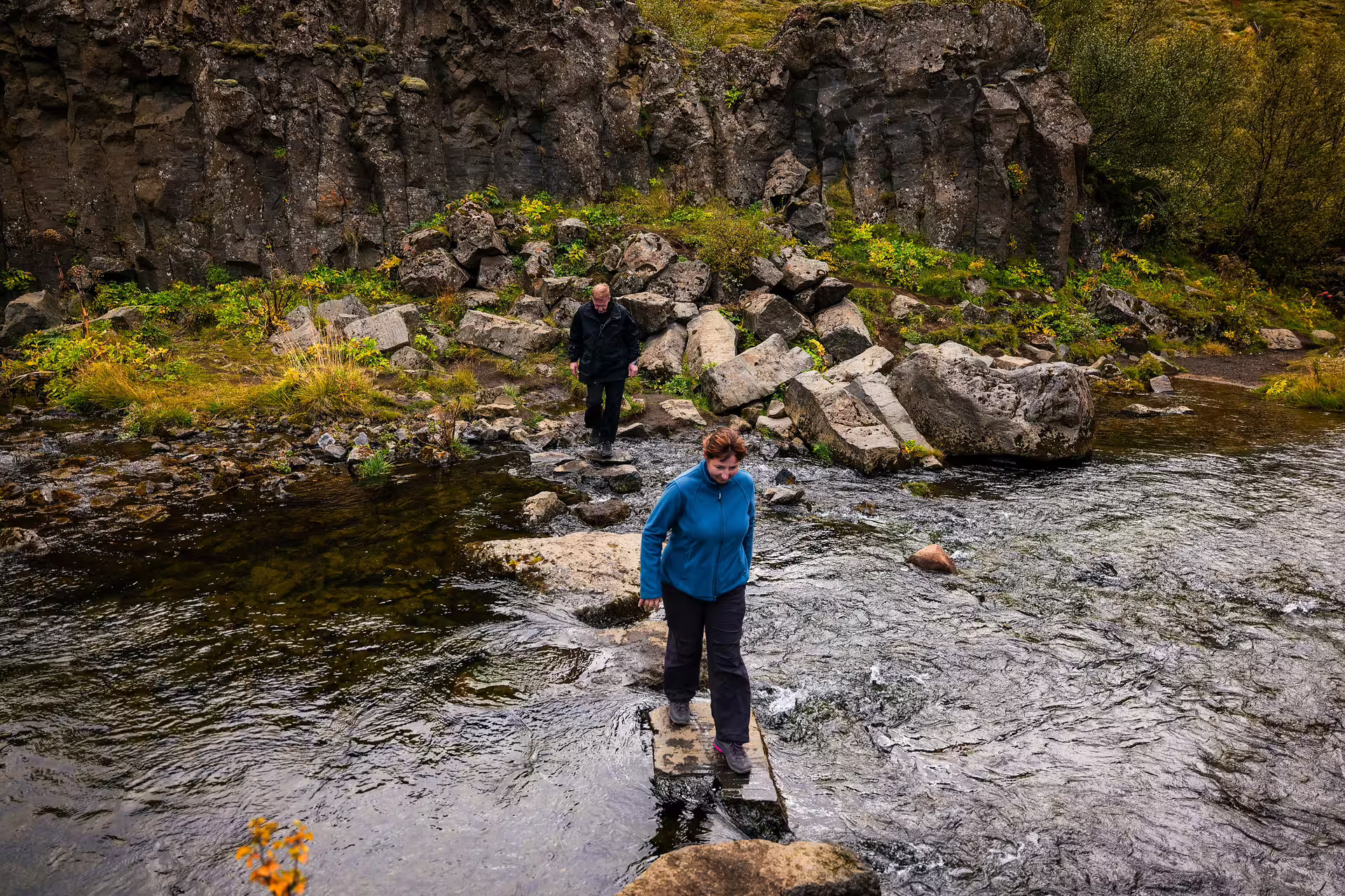 Two hikers carefully crossing a rocky highland river on a stone slab during The Hidden Highlands Iceland tour adventure