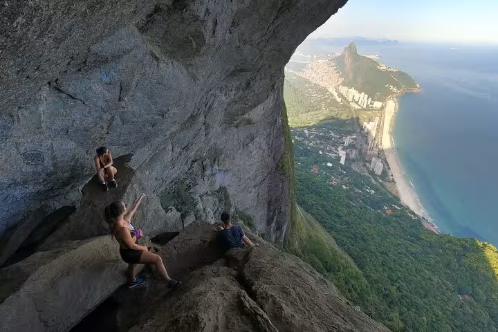 Group of hikers relaxes on Garganta do Céu ledge, overlooking stunning coastal views on Pedra da Gávea trail.