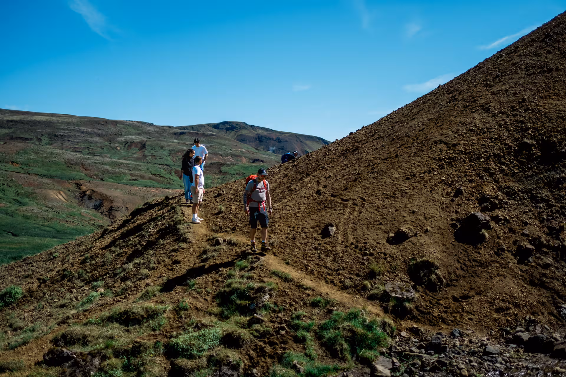 Hikers trekking along a narrow ridge in Iceland's geothermal valley near Hengill Volcano under blue sky