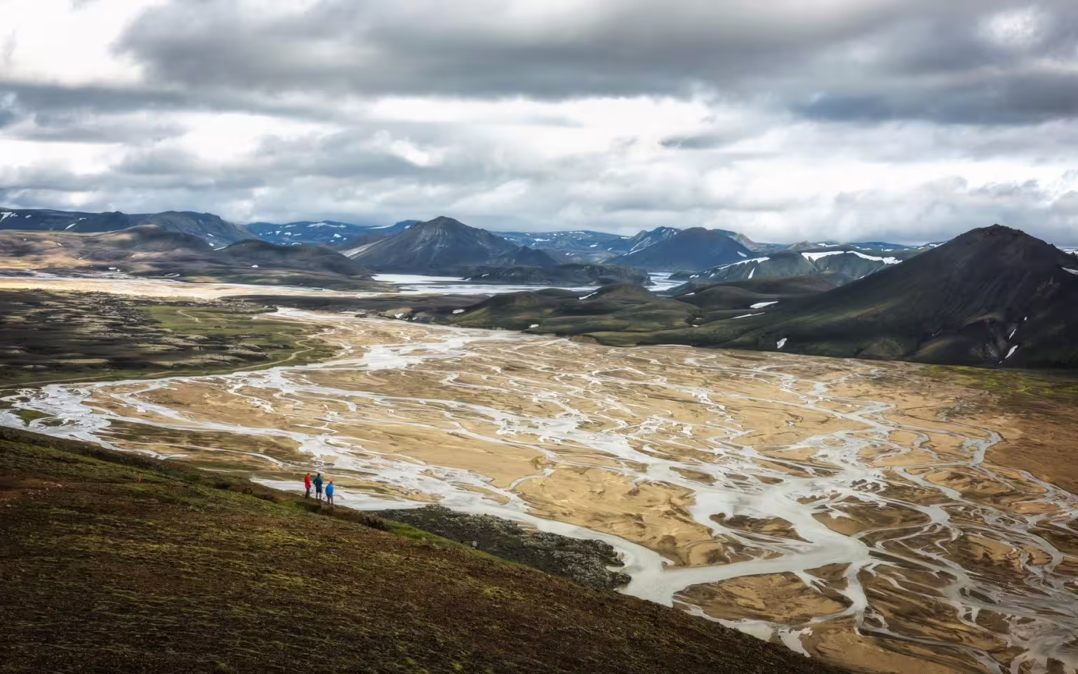 Hikers overlooking a vast braided glacial river valley with colorful mountains on Iceland grand tour adventure.