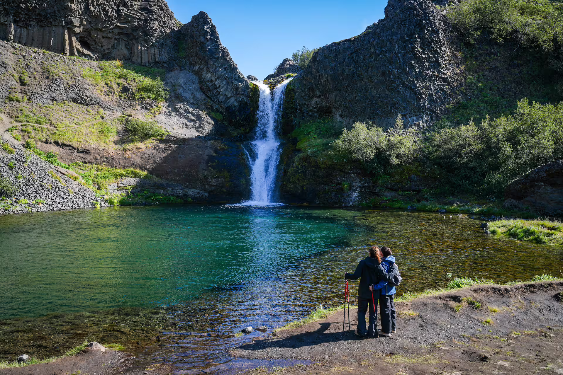 Two hikers with trekking poles admire a hidden Iceland waterfall cascading into a crystal-clear emerald pool in a rocky canyon
