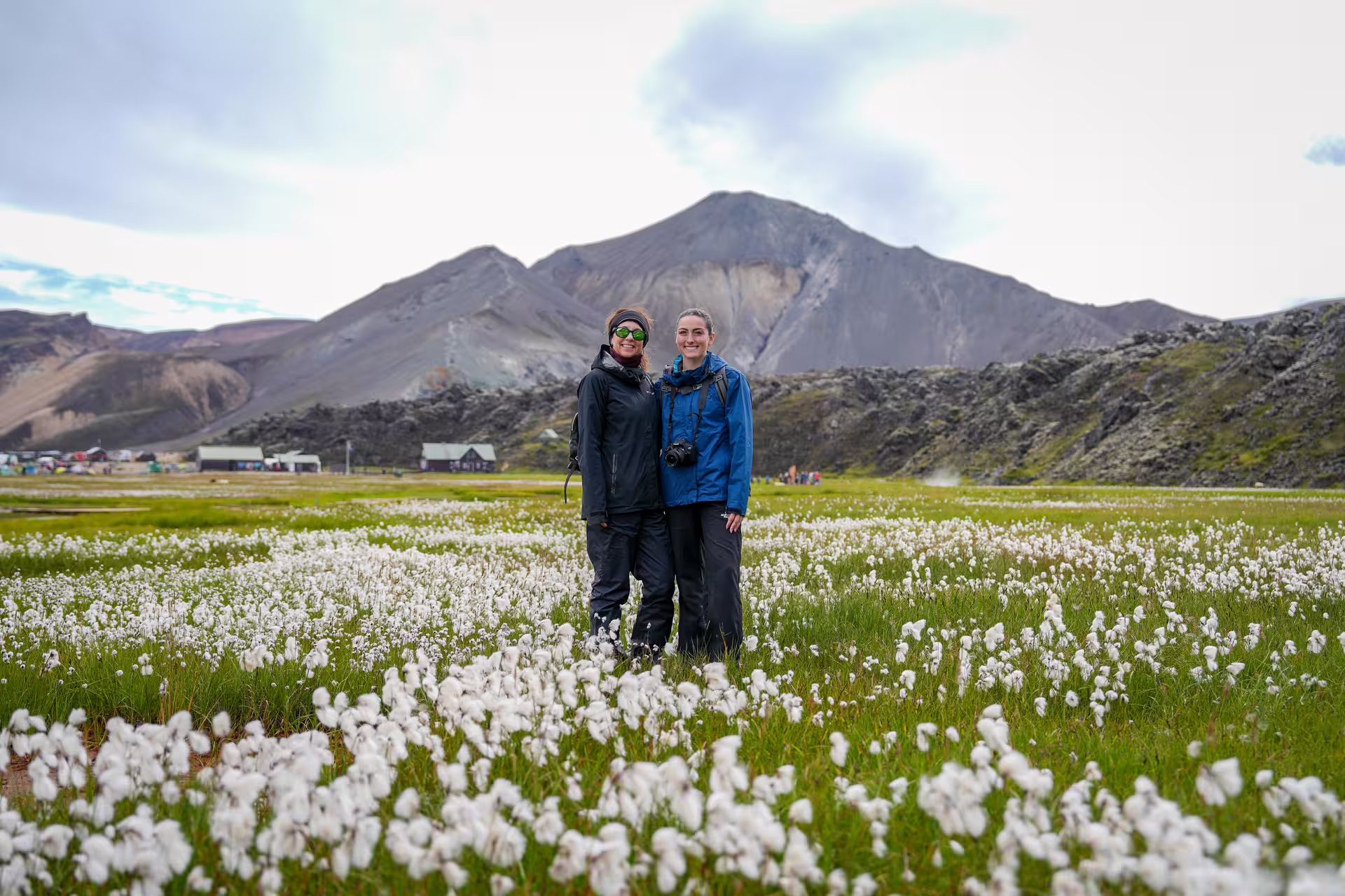 Two smiling hikers stand in a vast Iceland highland cotton grass field with dramatic volcanic mountains behind them.