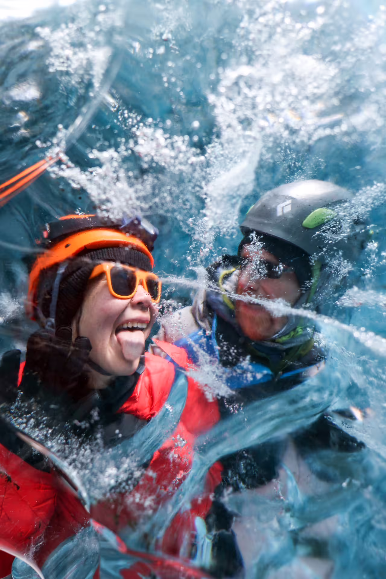 Two excited hikers wearing helmets laugh joyfully surrounded by sparkling blue glacier ice on an Iceland ice cave tour.