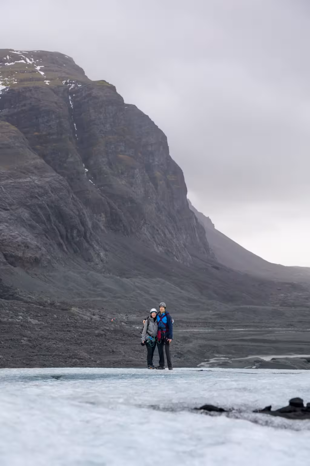 Two hikers with helmets and harnesses standing on an Iceland glacier with a dramatic rocky mountain backdrop