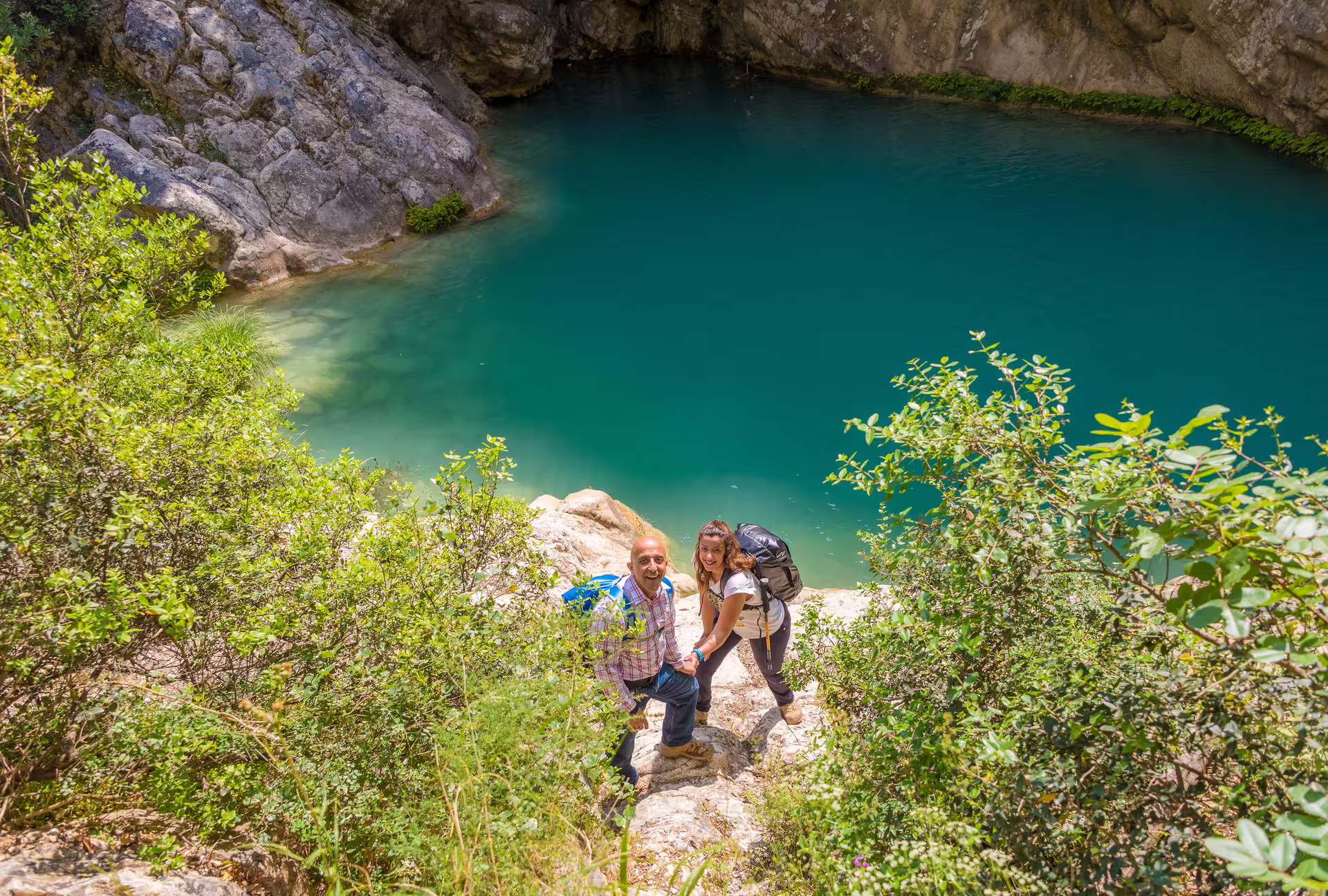 Hikers climbing rocky trail above turquoise pool on Polylimnio Waterfalls hike near Kalamata, Messinia, Greece