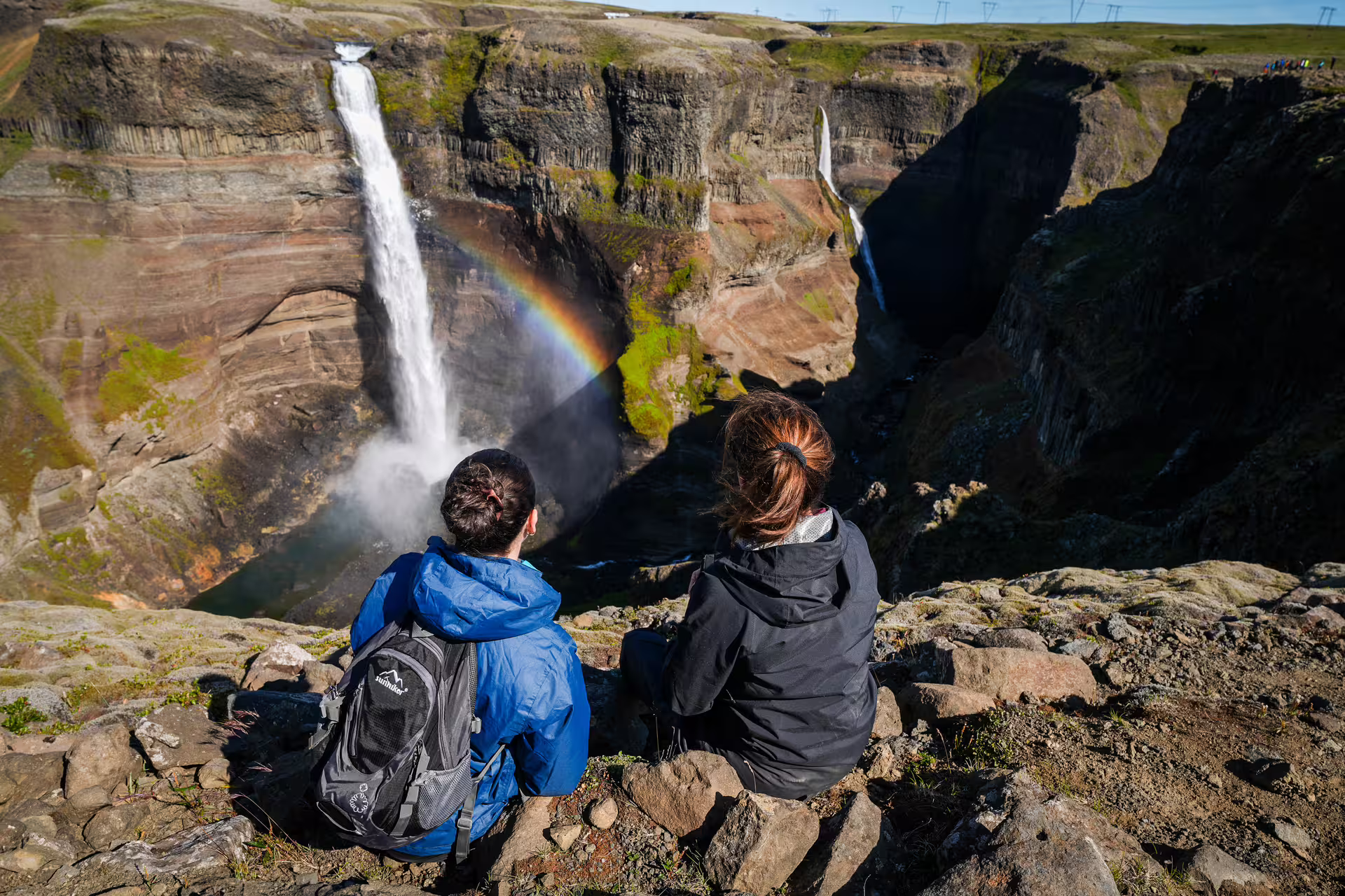 Two hikers sit on a cliff edge admiring a stunning rainbow over Haifoss waterfall on The Hidden Highlands Iceland tour