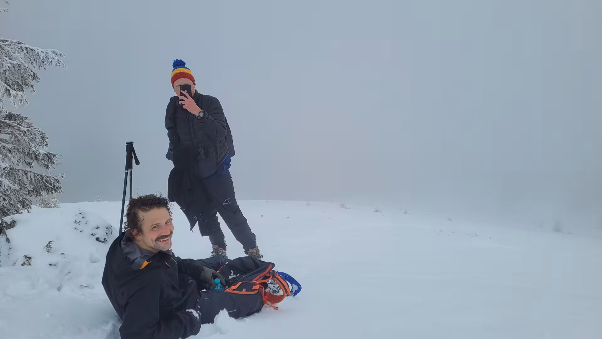 Two hikers resting in deep snow near snowy trees on Vladeasa Peak, Apuseni Mountains in winter fog