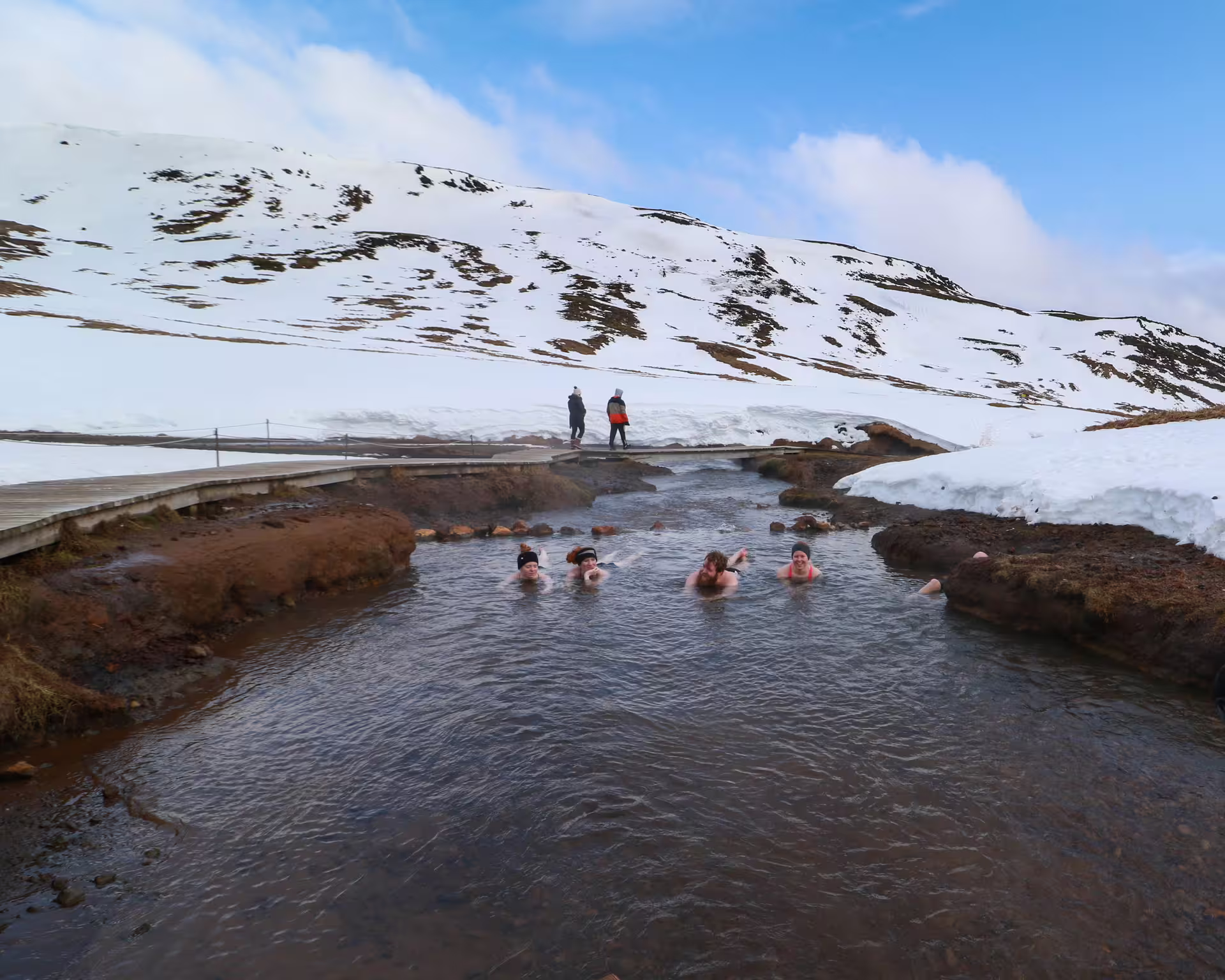 Hikers relaxing in the natural hot river of Reykjadalur Valley, surrounded by snow-covered hills and scenic Icelandic landscapes.