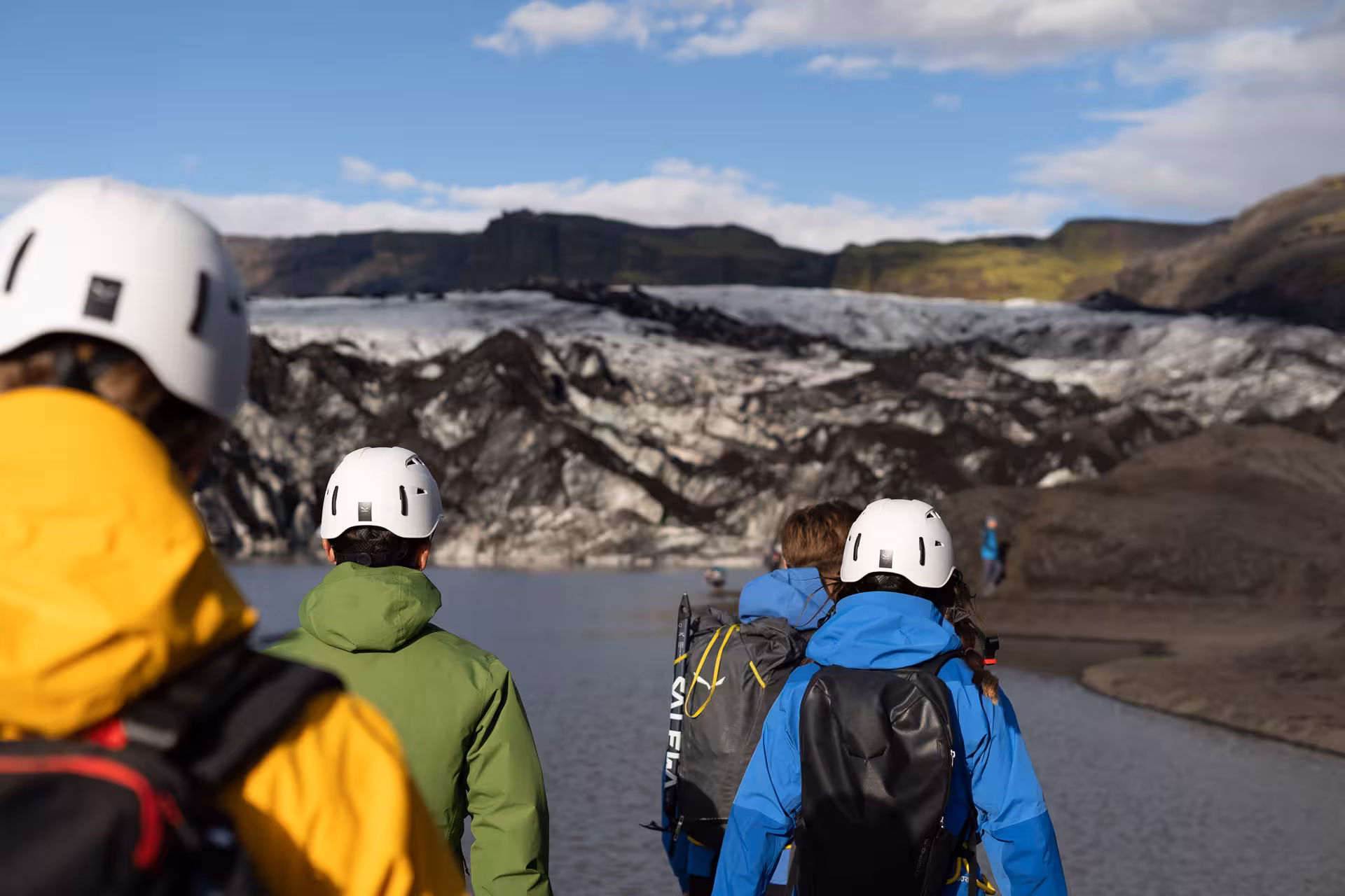Hikers in helmets walk toward Sólheimajökull glacier across the lagoon, starting a guided Iceland hike