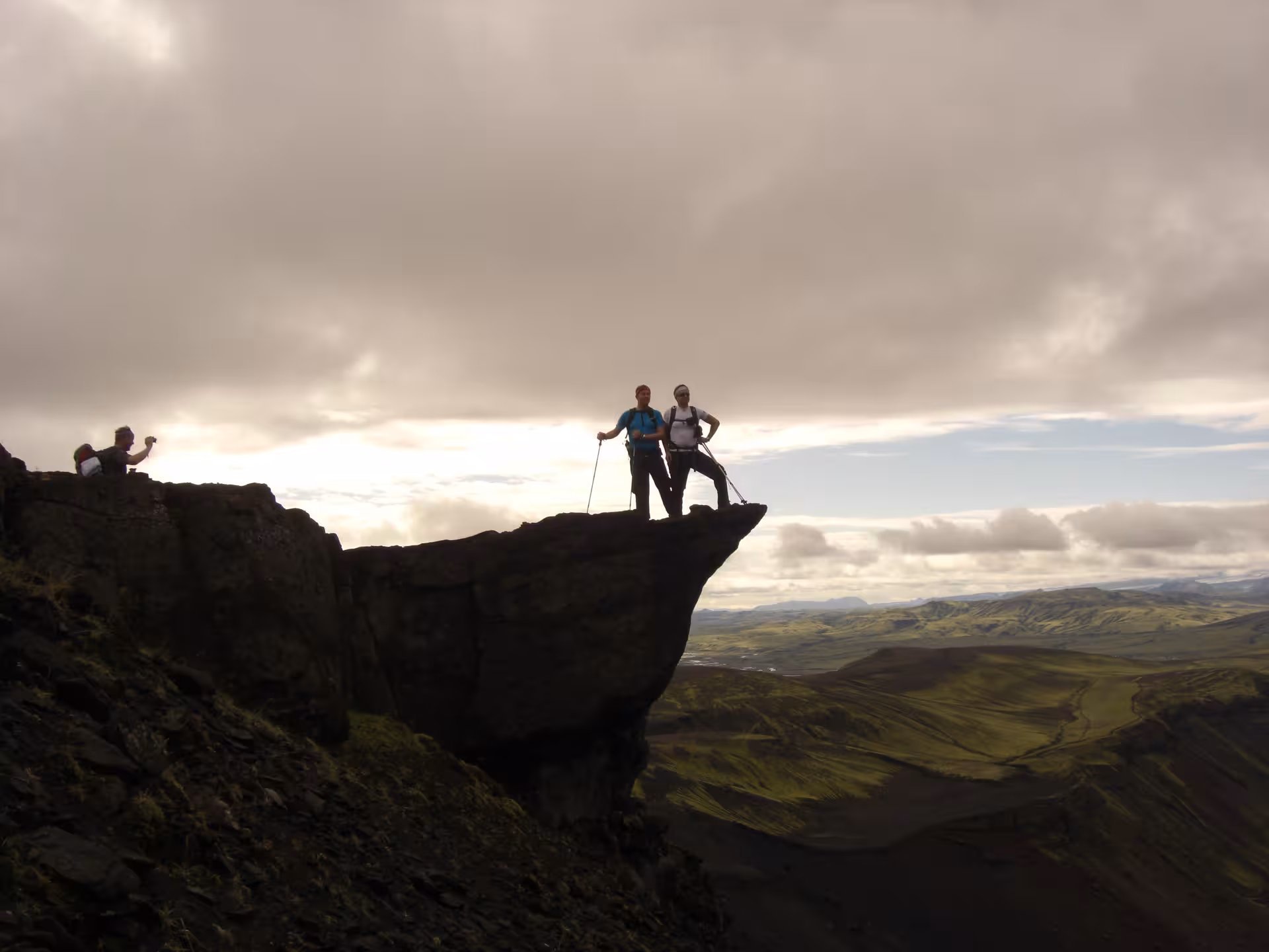 Two hikers standing on dramatic volcanic rock outcrop overlooking green Icelandic highlands valley