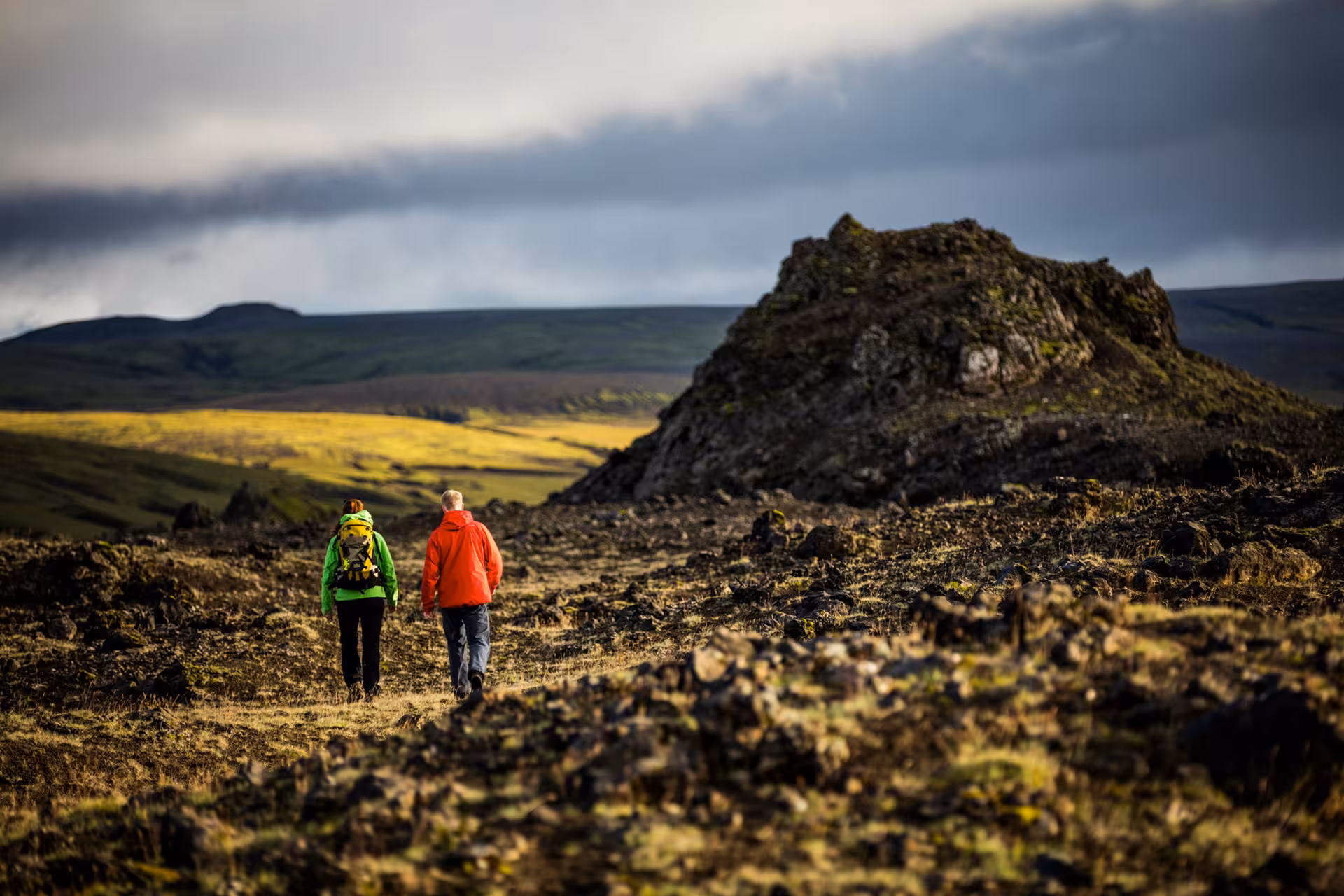 Two hikers walking through rugged volcanic lava field landscape in South Iceland under dramatic cloudy sky