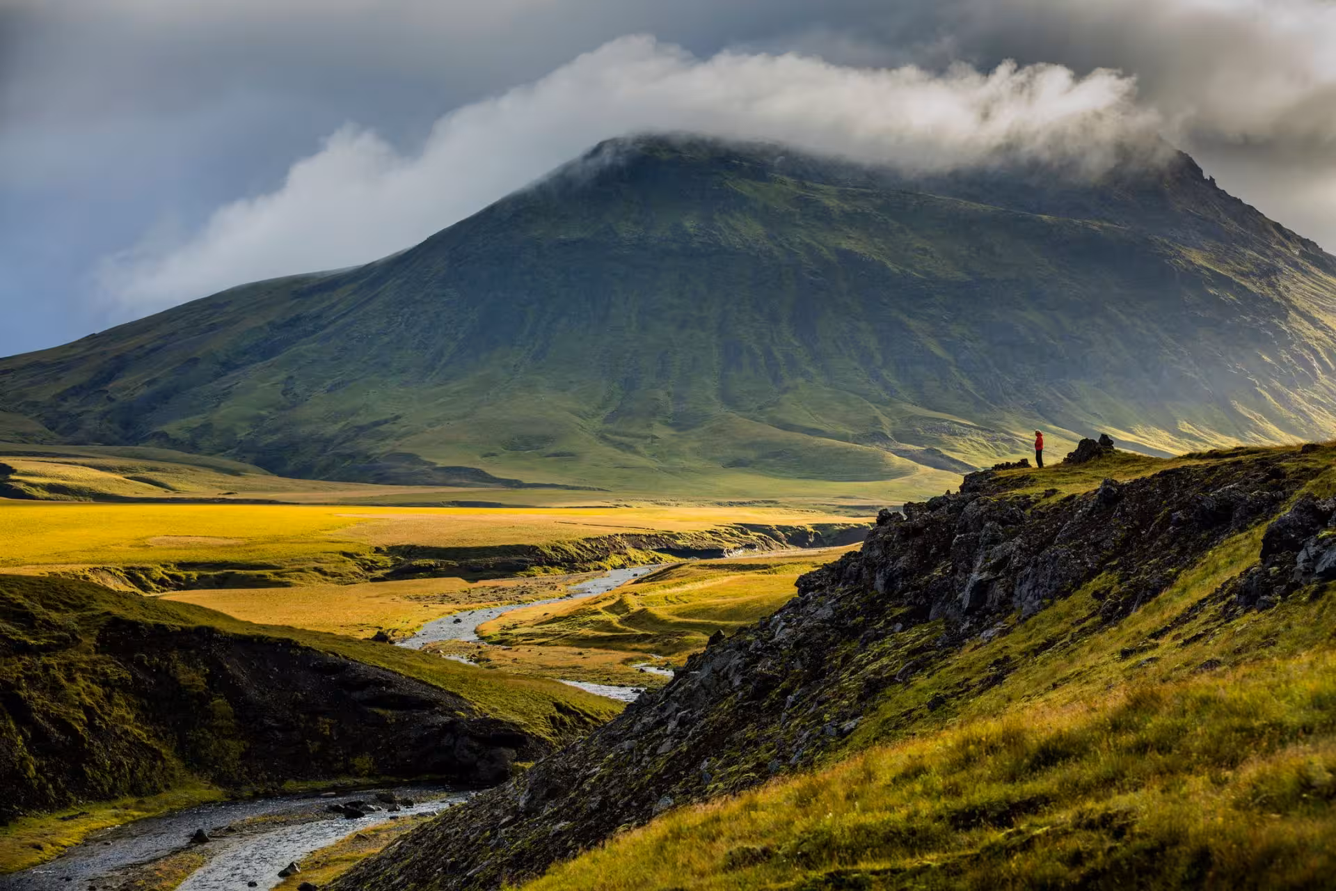 Hiker in red jacket standing on rocky ridge overlooking golden valley with green volcanic mountain and clouds
