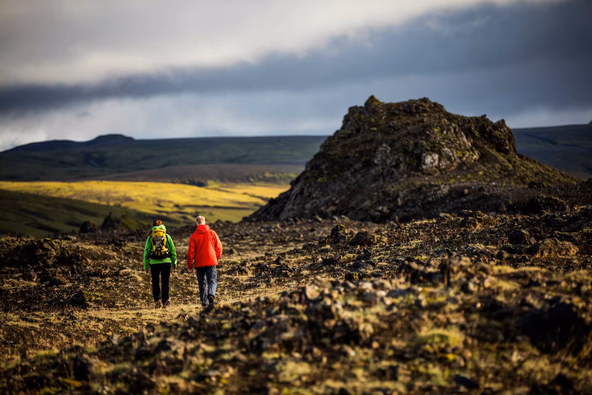 Two hikers in bright jackets trekking through dramatic volcanic landscape with rocky formations and rolling hills