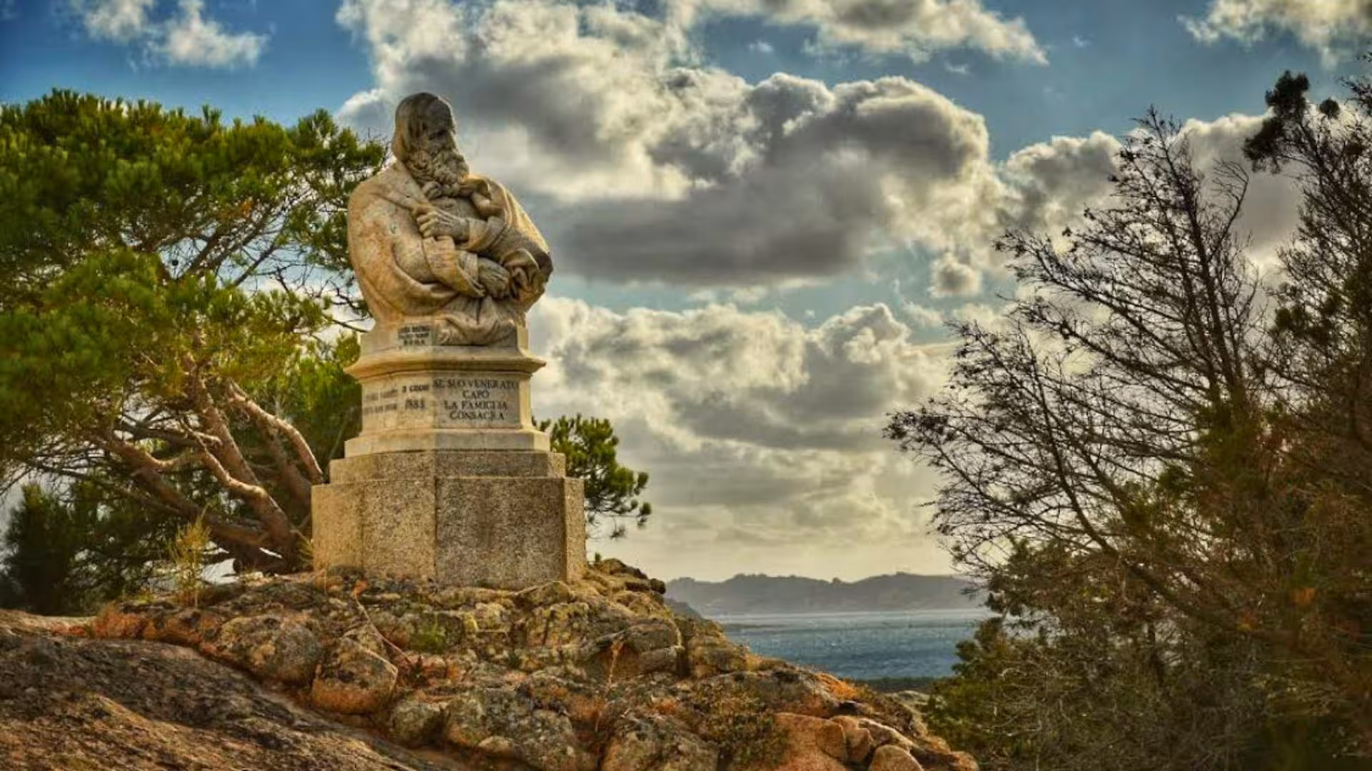 Historic statue surrounded by lush trees on a rocky hilltop in La Maddalena, overlooking a scenic coastal view.