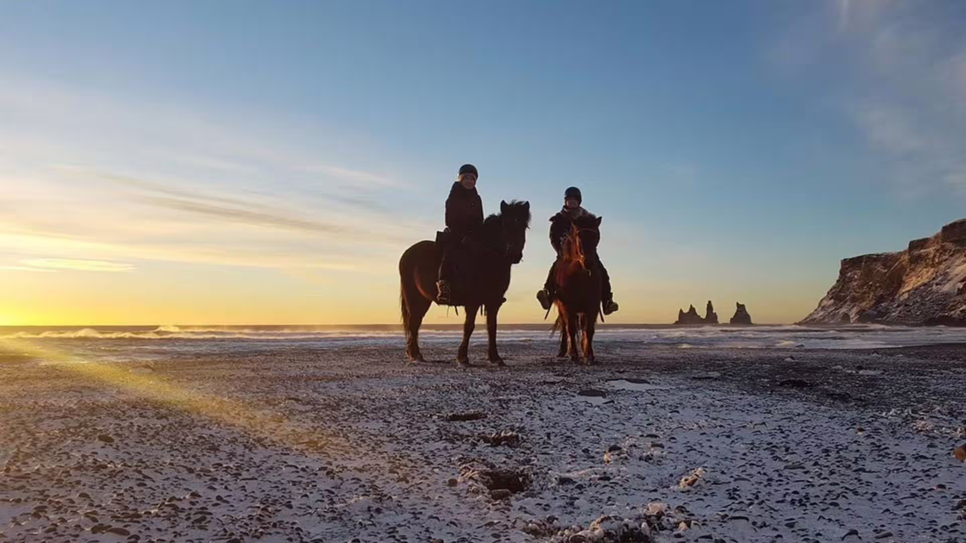Two riders on Icelandic horses on black sand beach at sunset with Reynisdrangar sea stacks in background
