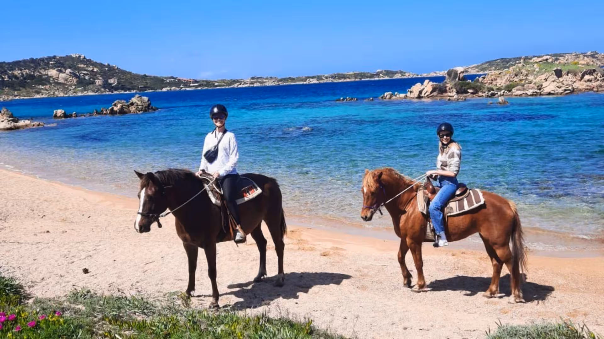 Two riders enjoy a horseback ride along the sandy beach in La Maddalena, with vibrant blue sea in the background.