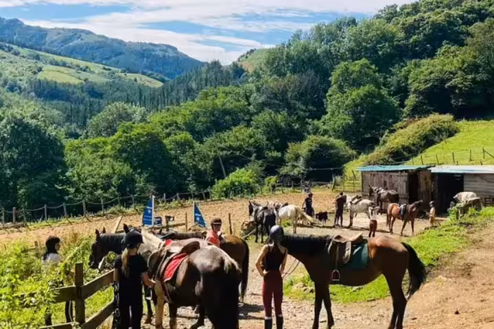 Riders preparing horses for a scenic horseback riding adventure amidst lush green mountains and clear blue skies.