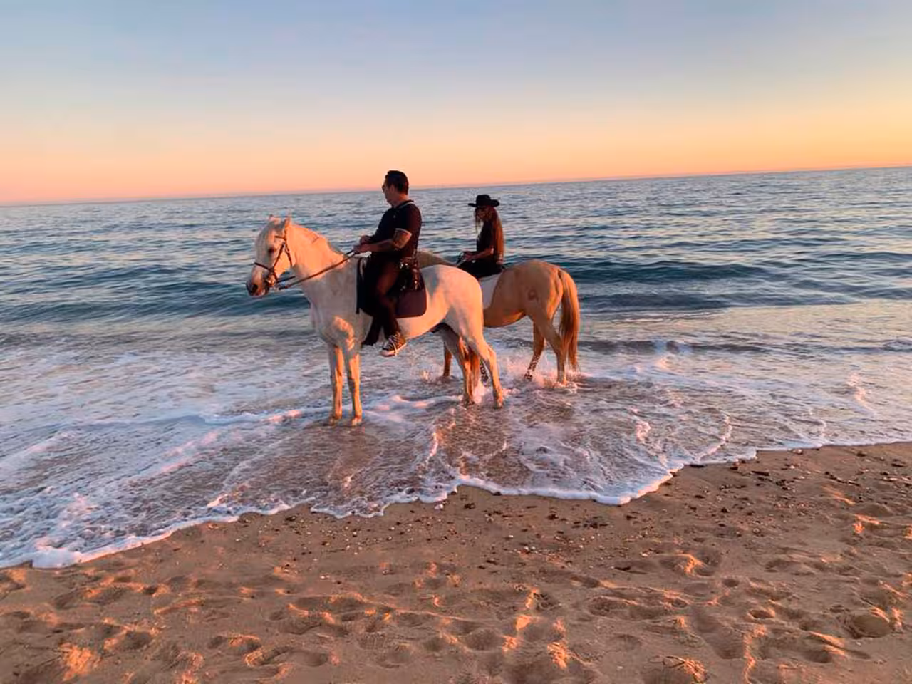 Riders on horseback enjoy the serene beauty of Quarteira's shoreline at sunrise, with waves gently touching the sand.