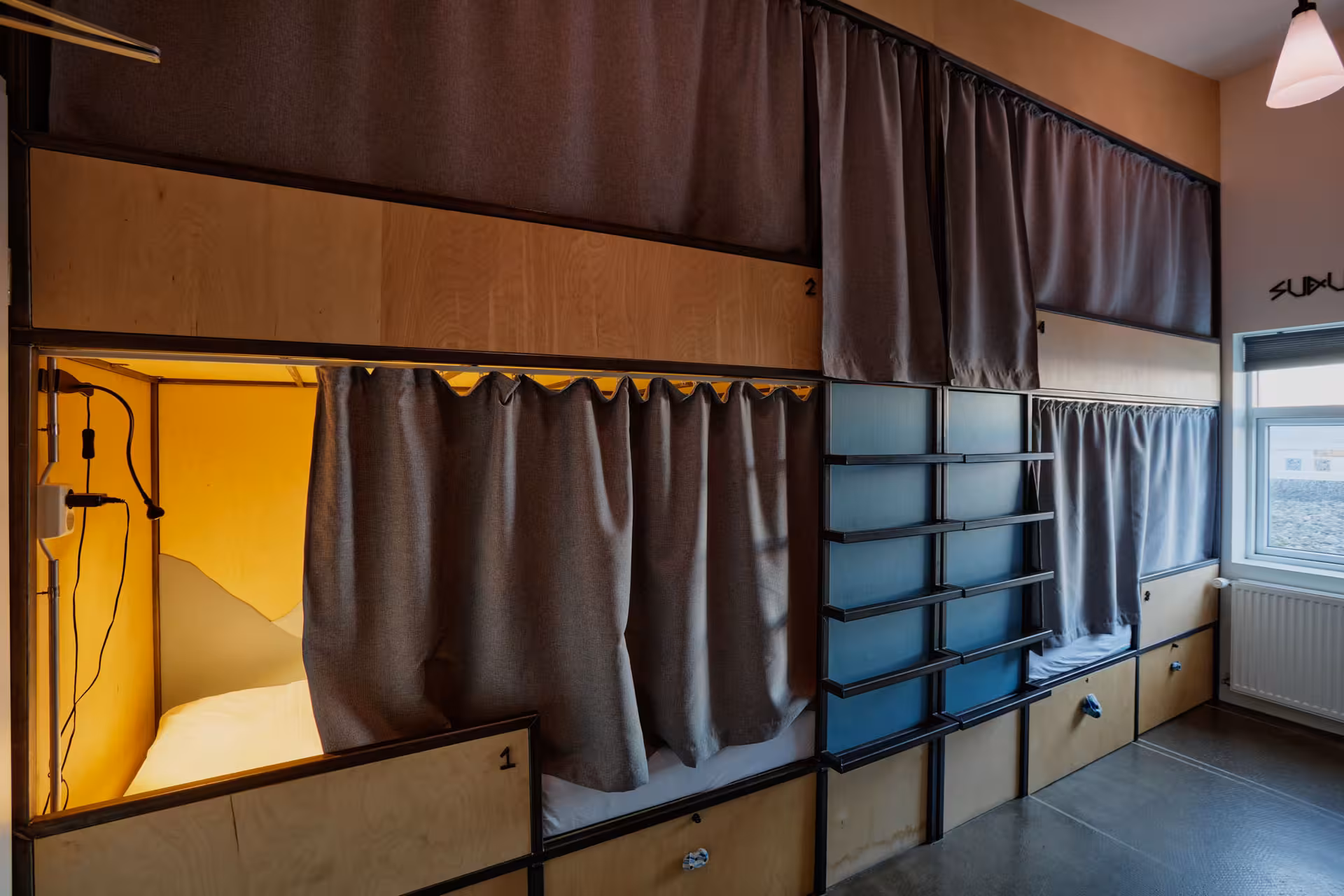 Pod-style bunk beds with privacy curtains and warm lighting in a hostel dormitory room