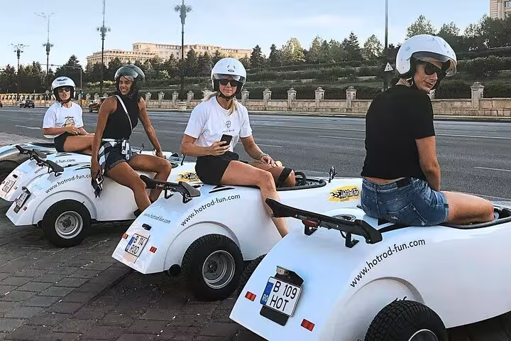 Four women wearing helmets riding white hot rod cars on a Bucharest city tour street