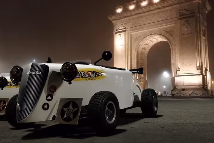 White hot rod car parked in front of the illuminated Triumphal Arch in Bucharest at night