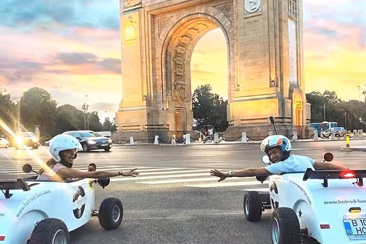 Two people riding white hot rod cars near the Arcul de Triumf in Bucharest at sunset