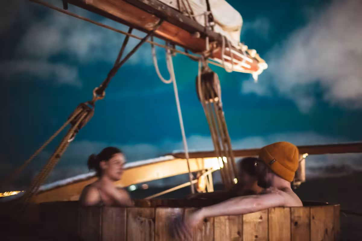 Guests relaxing in a steaming wooden hot tub on deck of a luxury cruise ship under the glowing Northern Lights arctic sky
