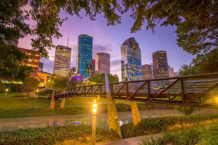 Houston downtown skyline at dusk with glowing skyscrapers, a wooden footbridge, and park greenery