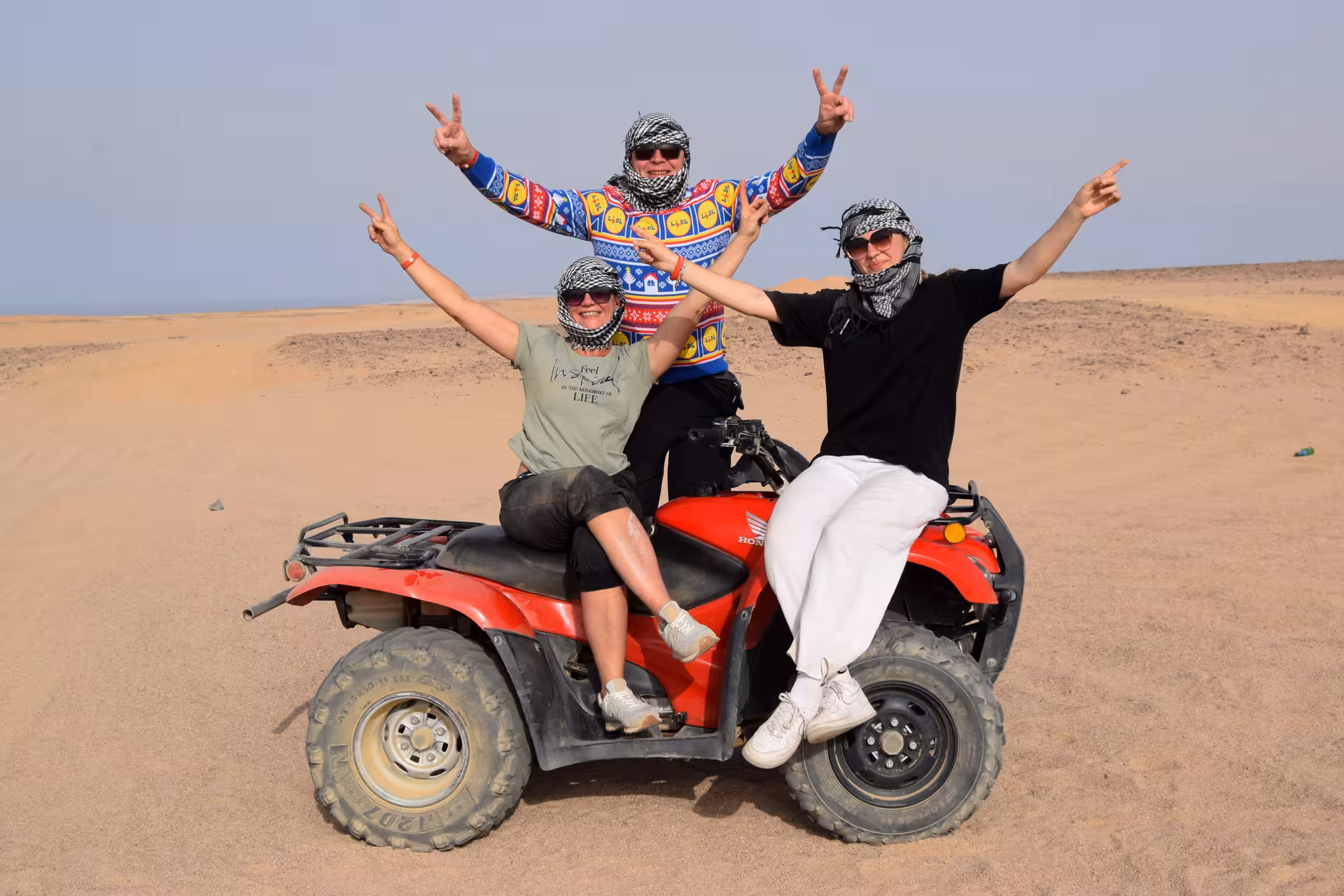 Friends posing on red ATV quad in Hurghada desert during quad safari tour with Bedouin village stop